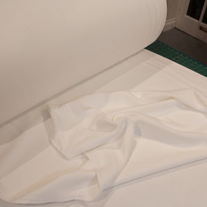 May include: A large roll of solid white fabric resting on a flat surface. The material is opaque and smooth in texture, with a portion of the fabric unrolled and draped in soft, loose folds across the foreground, showing its matte finish.