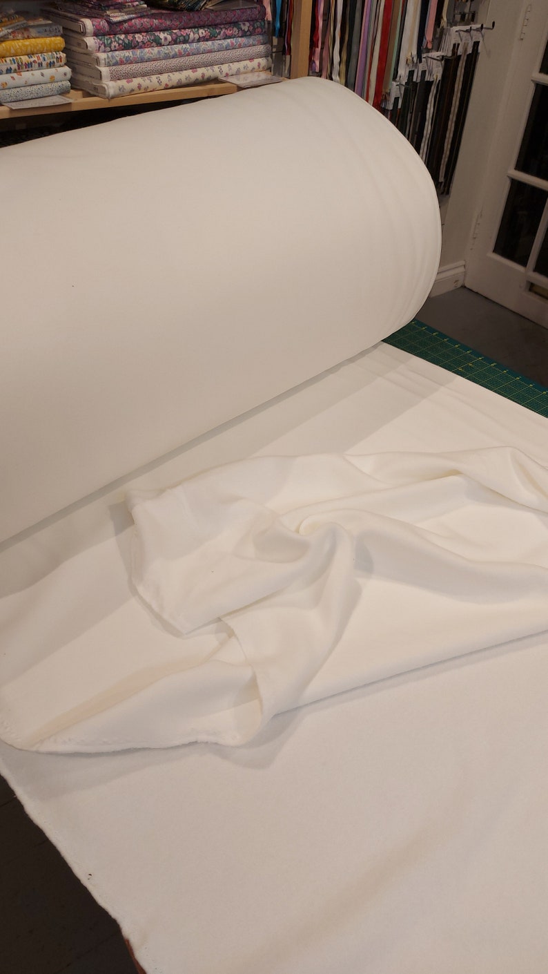 May include: A large roll of solid white fabric resting on a flat surface. The material is opaque and smooth in texture, with a portion of the fabric unrolled and draped in soft, loose folds across the foreground, showing its matte finish.