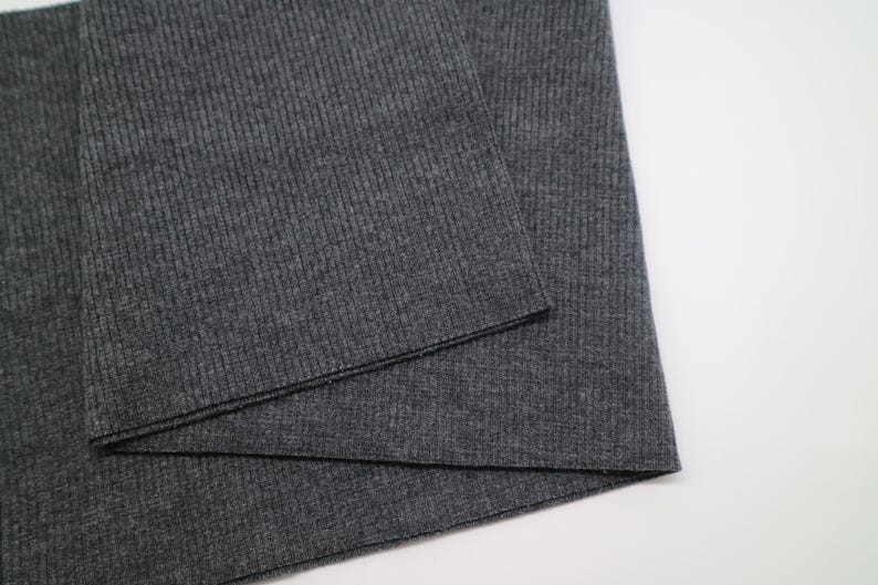 May include: A dark charcoal gray fabric featuring a fine vertical ribbed texture. The material is folded over itself, displaying a consistent knit pattern across the surface with a matte finish and a clean, straight edge along the fold line.