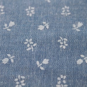 May include: Close-up of a blue fabric with a repeating pattern of small white floral designs. The fabric has a textured appearance, with the white flowers evenly spaced across the surface. The overall effect is a delicate, patterned textile.