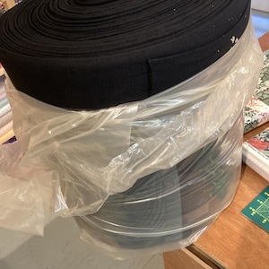 Può includere: Un grande rotolo circolare di nastro elastico nero, avvolto strettamente e fissato con un involucro di plastica trasparente. La superficie superiore del materiale nero presenta una consistenza a coste e un'etichetta bianca è fissata al centro.