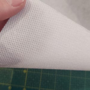 Puede incluir: Primer plano de una tela blanca perforada. La tela se levanta, revelando su textura y pequeños agujeros espaciados uniformemente. Una regla verde con marcas de centímetros es visible en primer plano.