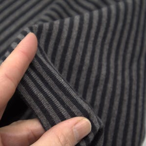 Pode incluir: Close-up de um tecido com listras verticais em tons de cinza e preto. O tecido está sendo segurado, mostrando sua textura e a direção das listras. O material parece macio e flexível.