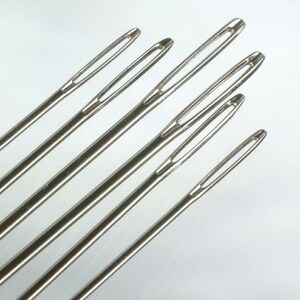 May include: A set of six silver-toned metal darning needles arranged diagonally. Each needle features a long, elongated eye designed for threading yarn or thick cord, with a smooth, polished finish and a sharp, tapered point at the opposite end.