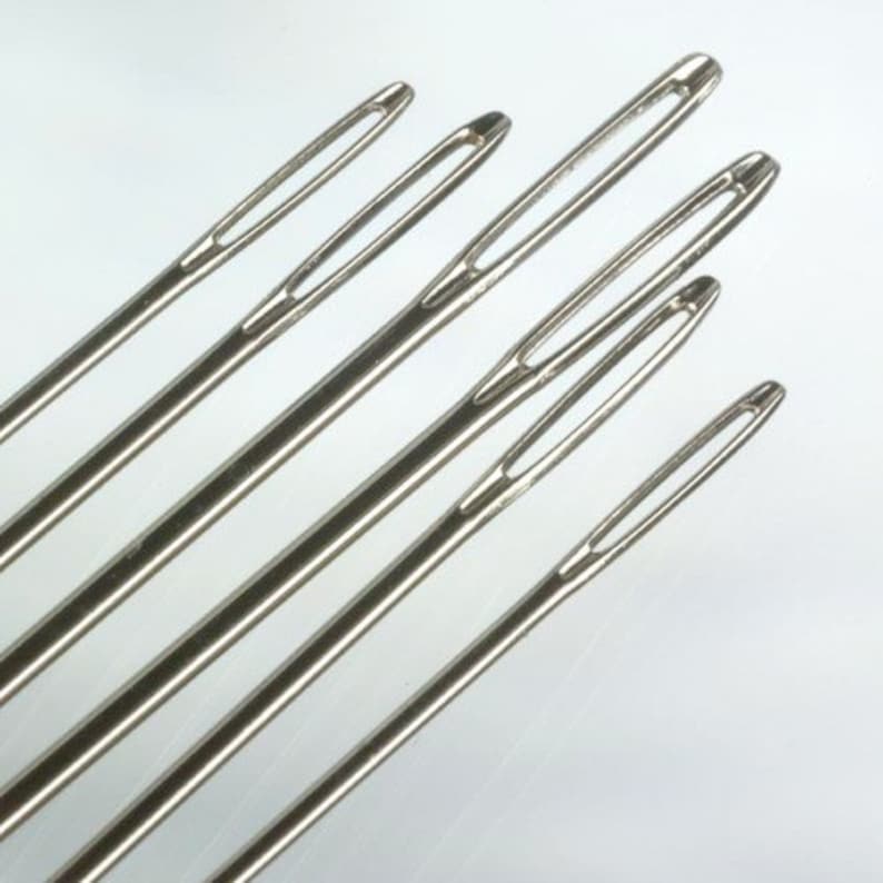 May include: A set of six silver-toned metal darning needles arranged diagonally. Each needle features a long, elongated eye designed for threading yarn or thick cord, with a smooth, polished finish and a sharp, tapered point at the opposite end.