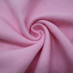 May include: A solid light pink fabric with a visible diagonal twill weave texture. The material is gathered into a central spiral swirl, creating soft folds and shadows that highlight the consistent pastel pink color across the textile surface.