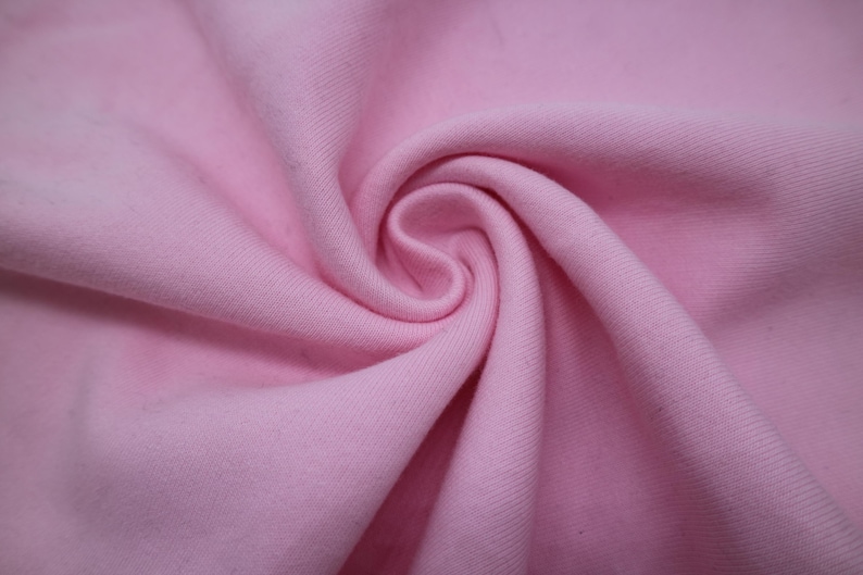 May include: A solid light pink fabric with a visible diagonal twill weave texture. The material is gathered into a central spiral swirl, creating soft folds and shadows that highlight the consistent pastel pink color across the textile surface.