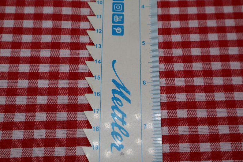 May include: A white ruler with blue markings and the word "Mettler" in blue script. The ruler is placed on top of a red and white gingham fabric.