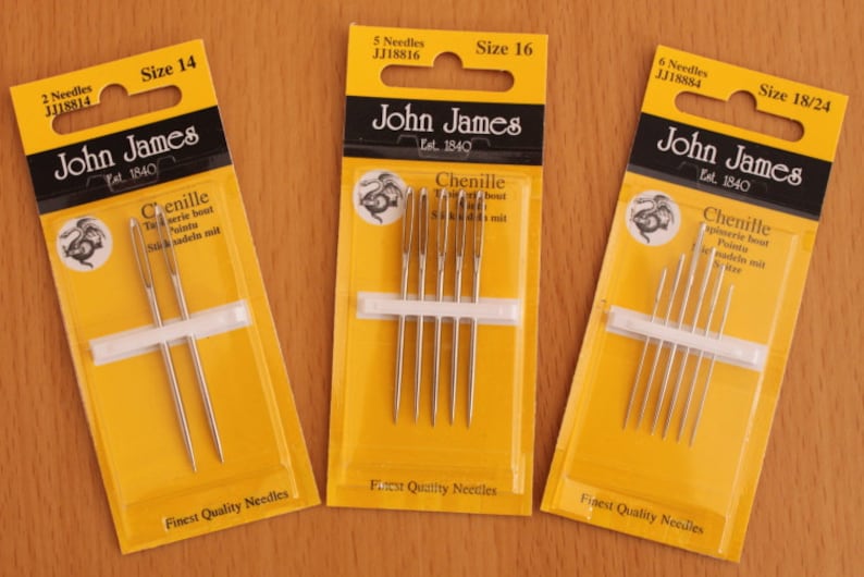 May include: Three yellow retail packages of John James Chenille needles. The left pack contains 2 size 14 needles, the centre pack contains 5 size 16 needles, and the right pack contains 6 size 18/24 needles. Each pack features a dragon logo.