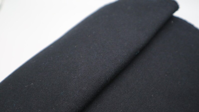 May include: A solid black fabric with a visible woven texture, featuring a distinct fold that creates a diagonal line across the surface. The material appears matte and opaque, showing a consistent dark color throughout the entire visible area.