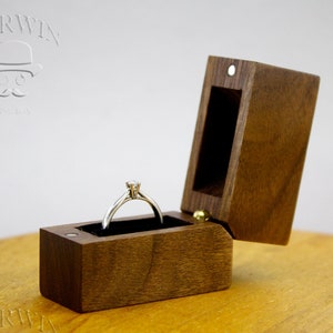 May include: A wooden ring box with a hinged lid, open to reveal a silver ring with a single diamond. The box is made of dark wood with a smooth finish.