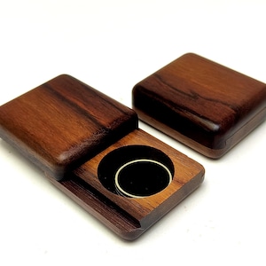 May include: Two rectangular dark brown wooden boxes with a smooth finish. One box is closed, while the other is open, revealing a circular cutout containing a simple gold-colored metal ring band resting inside a black interior lining.