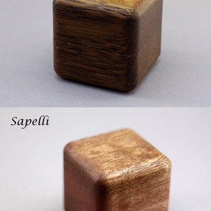 May include: Two wooden ring boxes, one made of Merbau wood and the other made of Sapelli wood. The boxes are both square and have a dark brown color. A silver ring is sitting next to the Sapelli wood box.