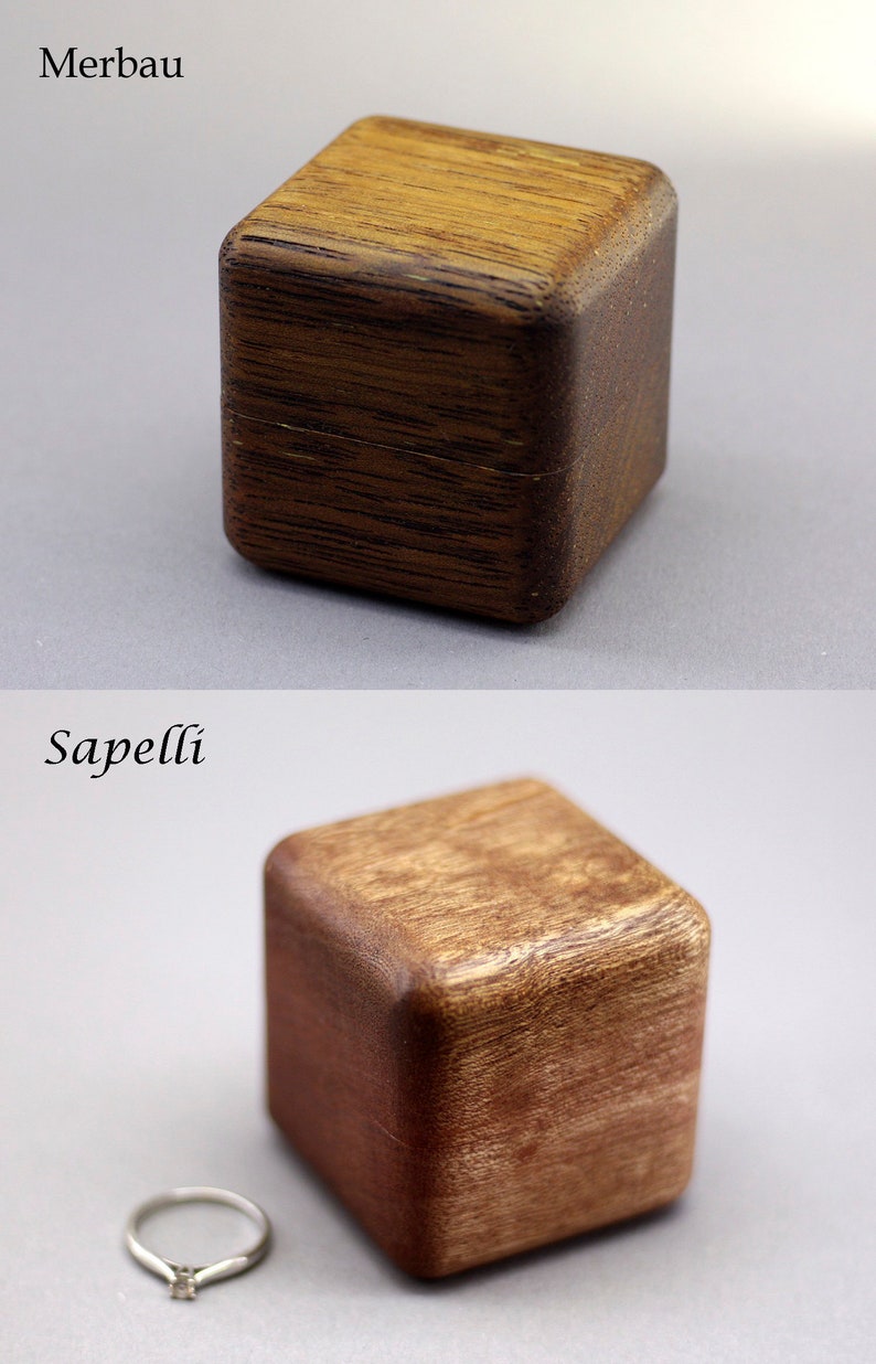 May include: Two wooden ring boxes, one made of Merbau wood and the other made of Sapelli wood. The boxes are both square and have a dark brown color. A silver ring is sitting next to the Sapelli wood box.