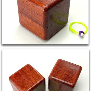 May include: Two small, square wooden boxes made of paduak wood. The boxes are closed and have a rich reddish-brown color. A green rubber band with a purple bead is in front of the boxes.