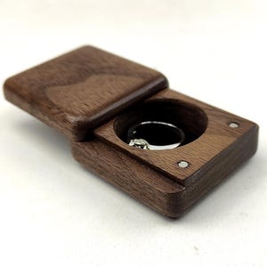 以下が含まれることがあります： スライド式の蓋が付いた長方形のダークブラウンの木製リングボックス。ボックスの内側には円形の切り込みがあり、小さな透明な石が付いたシルバーの指輪が収められています。ボックスの上面には、2つの小さな円形の金属製マグネットが見えます。