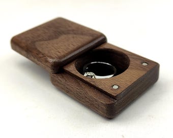May include: A rectangular dark brown wooden ring box with a sliding lid. The box features a circular interior cutout containing a silver ring with a small clear stone. Two small circular metal magnets are visible on the top surface of the box.