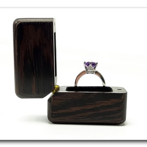 May include: A purple gemstone ring with a silver band sits in a dark wood ring box with the lid open.
