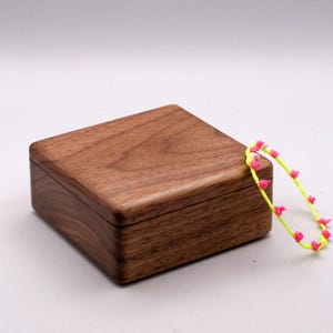 Könnte beinhalten: Eine Holzkiste mit Deckel aus Walnussholz. Die Kiste ist geschlossen und steht auf einer weißen Oberfläche. Ein leuchtend gelbes Armband mit rosa Akzenten liegt neben der Kiste.