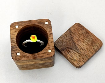 May include: A wooden ring box with a hinged lid. The box is made of dark wood with a natural grain pattern. The lid is open and a ring with a yellow and orange stone is inside.