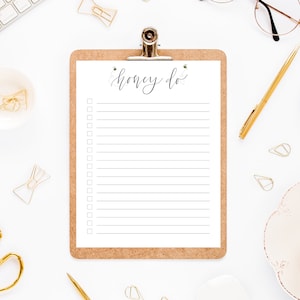 May include: A clipboard with a white sheet of paper with the words "Honey Do" written in black cursive. The clipboard has a cork backing and a gold clip. The clipboard is on a white surface with gold paper clips, a gold pen, a white cup with coffee, and a keyboard.