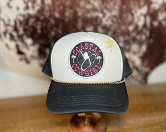 May include: White and black trucker hat with a black patch that says "Coastal Cowgirl" in pink and white lettering. The patch has a palm tree and a surfboard graphic. The hat has a gold chain trim.