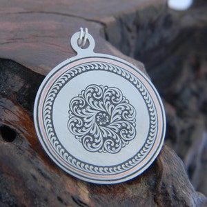 May include: A silver pendant with a detailed engraved design in the center, surrounded by a decorative border. The pendant has a rose gold rim and a loop at the top for attaching a chain.