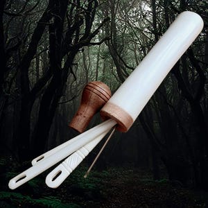 May include: A white cylindrical case with a wooden top and bottom, containing three white needles. The case is lying on a dark green and brown background.