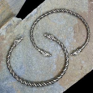 May include: Two silver-toned twisted wire bracelets with decorative bird head ends. The bracelets are displayed on a textured grey surface.