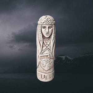 May include: A carved, off-white statue of a woman with a crown and long hair, holding a basket. The word "FULLA" is carved at the base. The statue is set against a dark, stormy background.