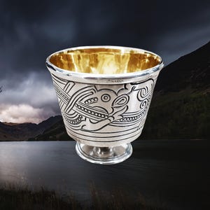 Könnte beinhalten: Ein silberner Trinkbecher mit goldenem Innenraum und einem komplizierten keltischen Knotenmuster. Der Becher hat einen runden Boden und eine breite, flache Schale.