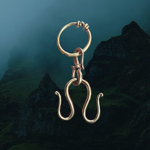 May include: A gold-coloured, ornate pendant with a circular top, a hook, and a stylised, symmetrical bottom design. The pendant is suspended against a blurred backdrop of dark mountains and a teal sky.