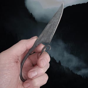 May include: A hand holding a small, black, forged knife with a curved handle. The knife has a sharp point and a slightly curved blade.