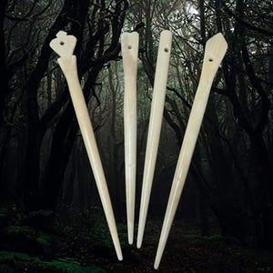 Puede incluir: Cuatro palos de pelo de hueso con diferentes formas en la parte superior. Los palos son todos blancos y tienen un acabado liso. El fondo es un bosque oscuro.