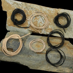 May include: Assortment of leather cord in various colors and sizes, including black, brown, and white. The cords are coiled and arranged on a stone surface.