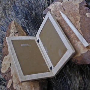 Könnte beinhalten: Eine Holzkiste mit zwei Scharnierklappen, jede mit einer braunen Wachsoberfläche. Die Kiste ist geöffnet und zeigt die Wachsoberflächen. Ein Holzgriffel liegt neben der Kiste.