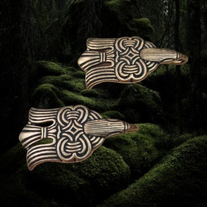 May include: Two bronze-colored, stylized bird-shaped brooches. Each brooch features intricate, symmetrical knotwork and linear designs. The background is a dark, mossy forest scene, enhancing the brooches' antique aesthetic.