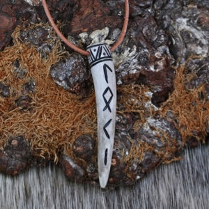 May include: A white antler pendant with black runes carved into it. The pendant is on a brown leather cord.