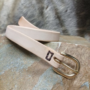 May include: A light brown leather belt with a gold buckle. The belt has a small square stitched into the leather.
