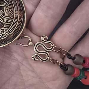 Pode incluir: Close-up de um fecho de colar dourado com um design de cobra. O colar apresenta contas vermelhas, verdes e castanhas. Uma porção de um pingente dourado é visível no canto superior esquerdo.