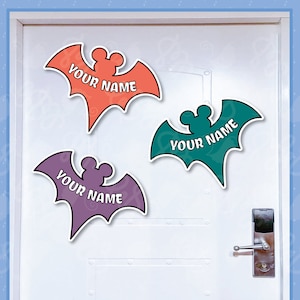 May include: Three Halloween door decorations shaped like bats with Mickey Mouse ears. The bats are in orange, teal, and purple with the text "YOUR NAME" on each.