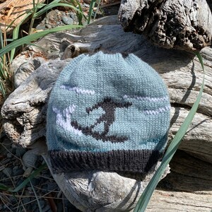 Könnte beinhalten: Eine blaue Strickmütze mit einem weißen und schwarzen Design einer fliegenden Möwe. Die Mütze hat einen schwarzen Rand.