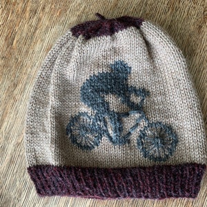 May include: A knitted beanie hat with a cyclist design. The hat features a light tan body with a dark burgundy brim and top. A blue and gray cyclist and bicycle are embroidered on the front.