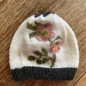 May include: White knitted beanie with a gray brim and embroidered pink flowers with yellow centers.