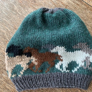 Puede incluir: Un gorro de punto verde, marrón, blanco y gris con un patrón de caballos corriendo.