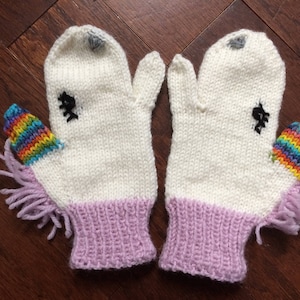 Op de afbeelding: Een paar witte gebreide wanten met paarse manchetten en regenboog garen eenhoornhoorns.