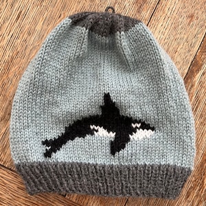 May include: A gray and black knitted hat with a black and white orca design. The hat has a gray brim and a black top.