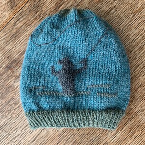 May include: A blue knitted hat with a dark gray fisherman design. The hat has a green brim.