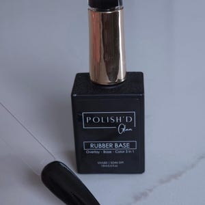 May include: A black square nail polish bottle with a gold cap and the text "POLISH'D Glam RUBBER BASE". A clear plastic nail swatch displays a glossy black nail polish color.
