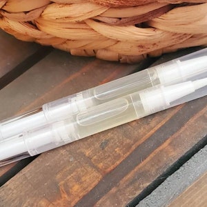 May include: Three clear nail oil pens with white tips and caps are arranged on a wooden surface. The pens are filled with a light yellow liquid. A woven basket is in the background.